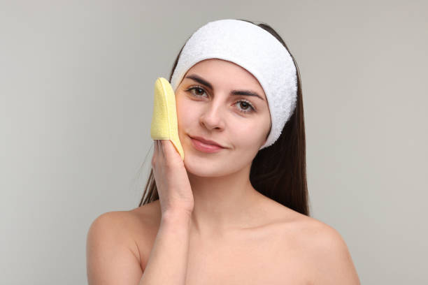 istock Young woman with headband washing her face using sponge on light grey background 2202967719