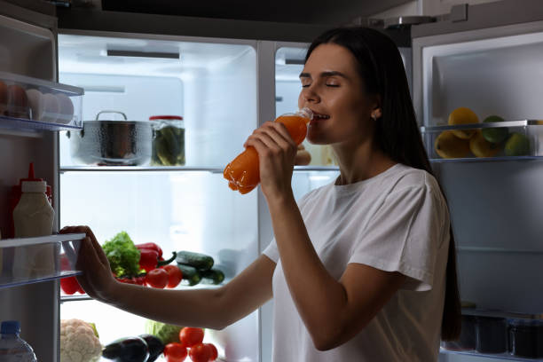 istock Young woman drinking juice near modern refrigerator at night 2202969588
