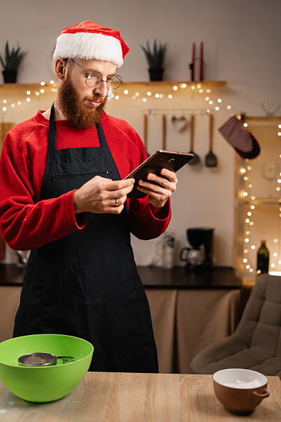 istock Bearded Man in Santa hat making dough while watching online cooking course via tablet in decorated kitchen on Christmas holiday 2202977809
