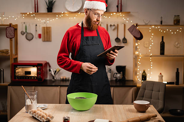 istock Man in Santa hat preparing dough for baking while reading recipe from tablet, standing in Christmas-decorated kitchen, surrounded by ingredients and utensils 2202977815
