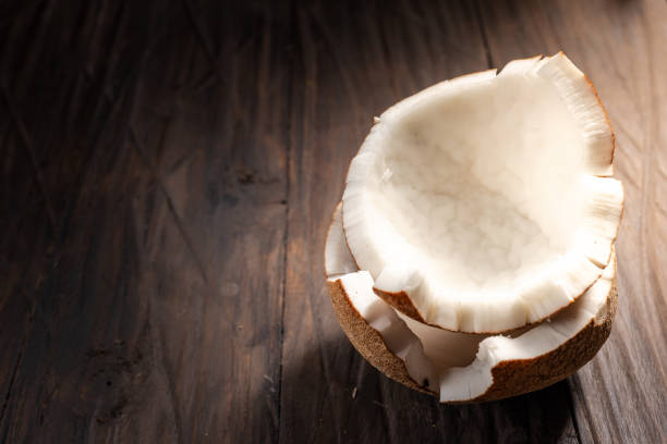 A cracked coconut shell on a rustic wooden table. A key ingredient in Indonesian cuisine, often used in Eid dishes like opor ayam. The rough brown husk contrasts with the smooth white flesh inside