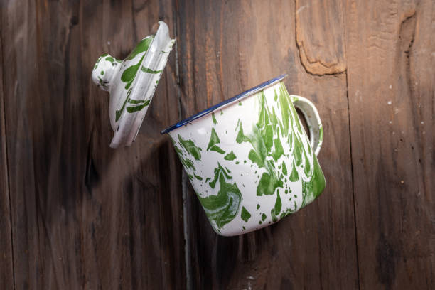 A vintage enamel mug with a green marble pattern and a detached lid, placed on a rustic wooden table. A classic Indonesian kitchen item often used during Eid gatherings for serving traditional drinks