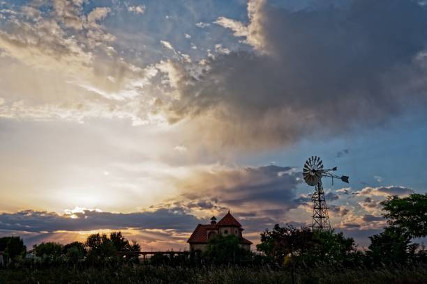 A serene rural landscape at sunset featuring a silhouette of a windmill and a house against a dramatic sky.