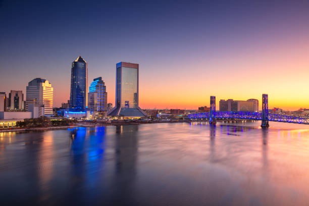 Jacksonville's skyline at sunrise with modern skyscrapers reflecting in calm water. Tall, glass-covered buildings, including a pyramid-topped structure, are illuminated with blue and white lights. A distinctive blue-lit bridge spans a wide river on the right, contributing to the vibrant urban landscape. The sky transitions from deep blue to warm orange hues, highlighting the serene water surface. Palms and low vegetation are visible near the waterfront, adding to the scenic view of an urban coastal setting.