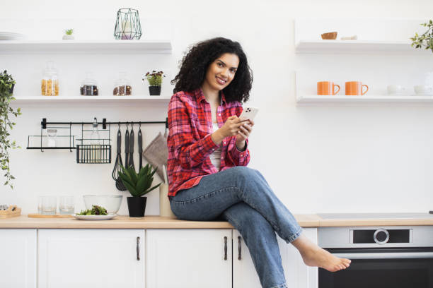 istock Smiling young woman using smartphone sitting on modern kitchen countertop 2203006504