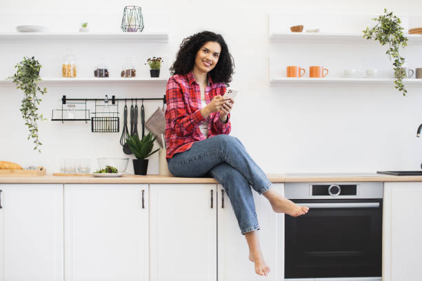 istock Smiling woman using smartphone sitting on kitchen counter in modern apartment 2203006515