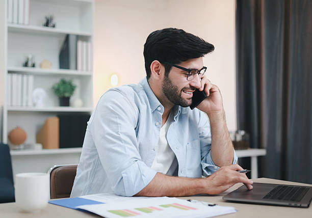 istock A man working on a laptop at home with a happy smile, using a phone to explain his work. 2203006568