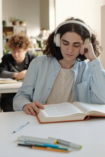 istock Student at Desk in College Library Reading Book With Headphones 2203010575