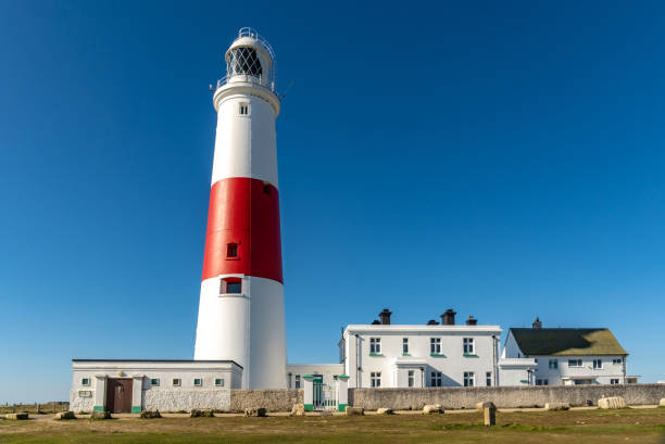 A striking stone lighthouse stands along a breakwater, illuminated by soft sunlight against a clear blue sky. Surrounded by calm ocean water, this peaceful coastal scene captures the timeless beauty and purpose of maritime architecture. Ideal for travel, nautical, and landscape themes.