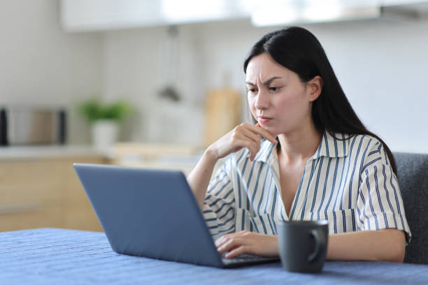 Suspicious asian woman using laptop in the kitchen stock photo