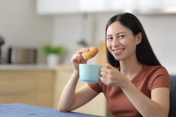istock Happy asian woman eats doughnut drinks coffee and looks at you 2203021017