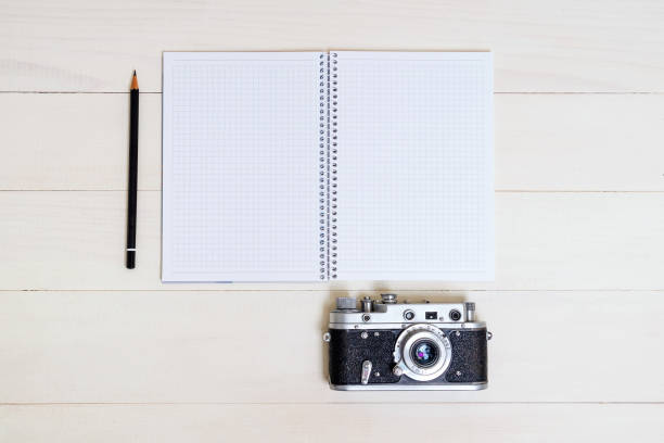 Photo camera, pencil and spiral notebook for writing down ideas, flat lay on a light wooden table.