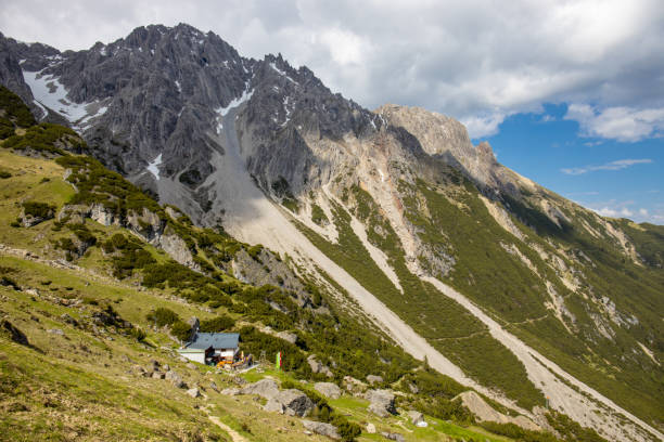 A mountain hut situated on a green hillside with rugged peaks on background. Flags are displayed near the building. The landscape includes rocks, grass, hutte mountain Refugio in Austria neat Imst, Muttekopfhütte