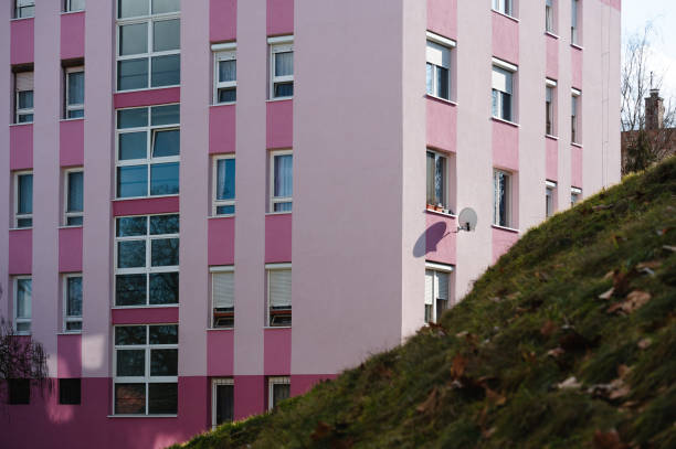 A modern pink apartment building with large windows and balconies, partially obscured by a grassy hill in the foreground. The contrast between the vibrant architecture and the natural landscape creates a visually appealing urban scene.