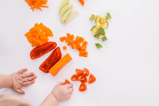 istock A child's hands reaching for freshly chopped vegetables on a white surface. Includes diced avocado, carrot, apple slices, bell pepper, and tomato pieces. Minimalist food composition. 2203051885