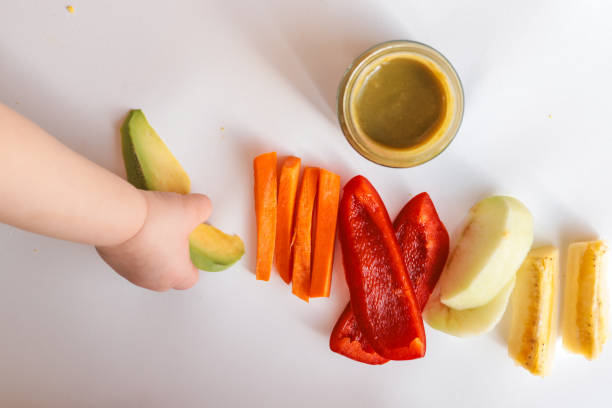 A child's hands reaching for freshly chopped vegetables on a white surface. Includes diced avocado, carrot, apple slices, bell pepper, and tomato pieces. Minimalist food composition.