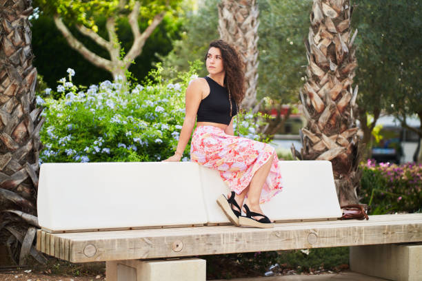 a stylish young woman in a hat sunglasses and a black dress is sitting on a bench in front of the house on a summer day