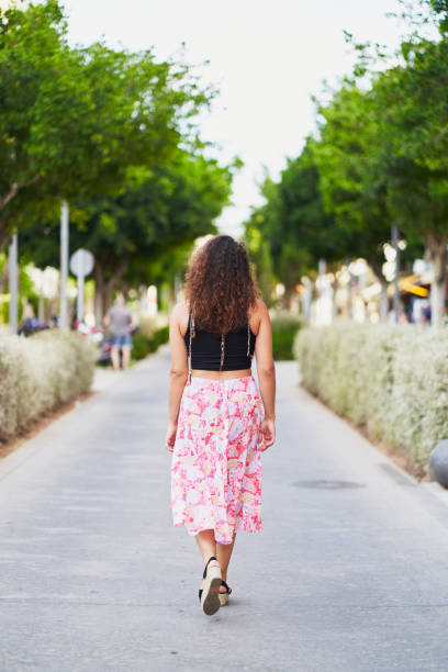 Young woman with long, curly brown hair, wearing a black top and a long pink floral skirt, walks away from the camera down a paved street lined with trees and bushes, enjoying a leisurely stroll