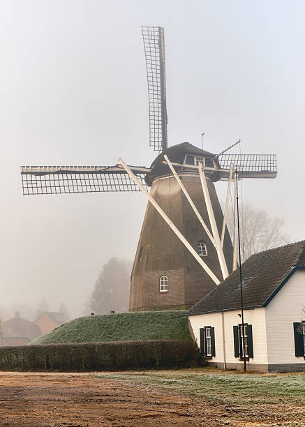 Atmospheric View on Traditional Windmill Keetmolen in Ede, Netherlands, with old house in front, rising up through winter mist.