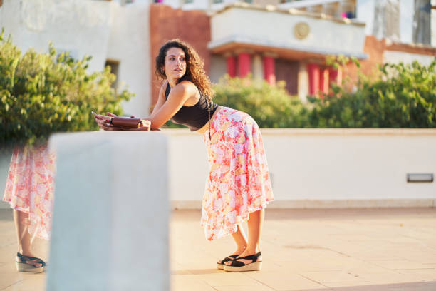 Young woman leaning on a balcony, holding her smartphone and purse, soaking in the stunning views of a mediterranean town, radiating happiness and relaxation under the sun