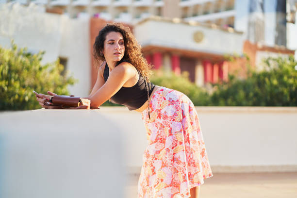 Young woman with curly hair, wearing a black top and floral skirt, leans on a wall while using her mobile phone, with a blurred urban background