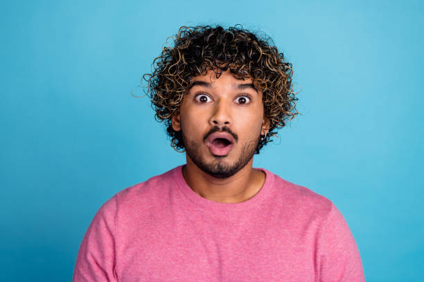 istock Surprised young man with curly hair against a blue background wearing a pink pullover 2203059881