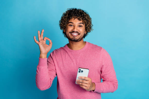 istock Happy Young Man Gesturing OK Sign and Holding Smartphone Against Blue Background While Smiling in Casual Pink Outfit 2203059957