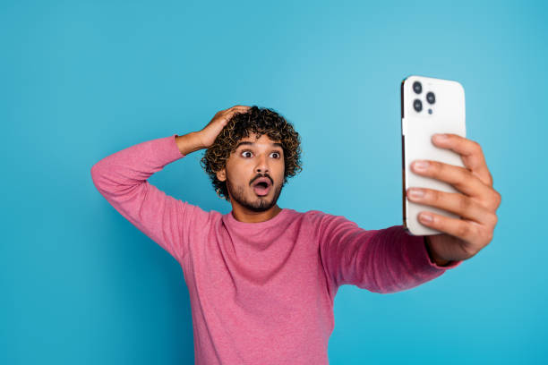 istock Young man in a pink pullover reacting with surprise while taking a photo on his smartphone against a blue background 2203059968