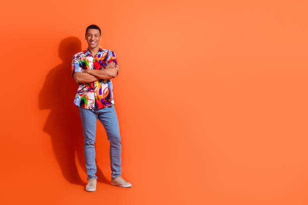 istock Confident young man in stylish printed shirt standing against a vibrant orange backdrop, expressing a cheerful and casual vibe 2203060603