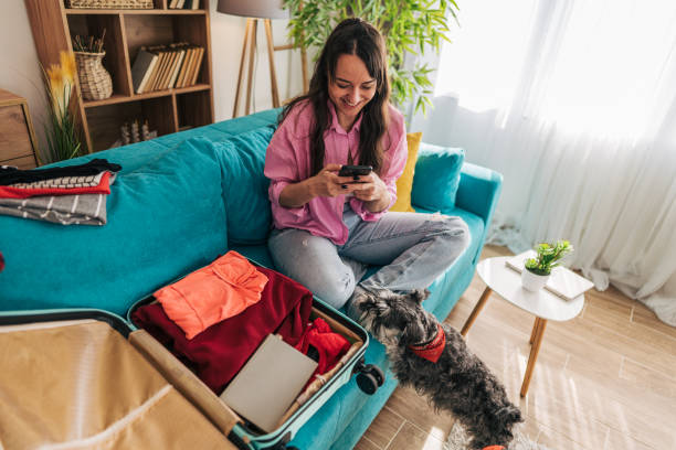 istock Young woman using smartphone while packing suitcase at home with pet 2203064235