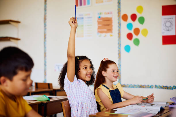 istock Elementary schoolgirl with hand raised in class, answering a question 2203067305