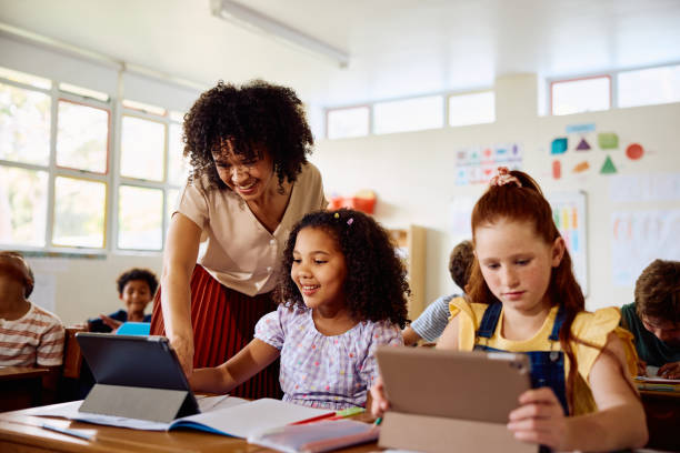 Happy young female teacher talking to schoolgirl using digital tablet in class stock photo