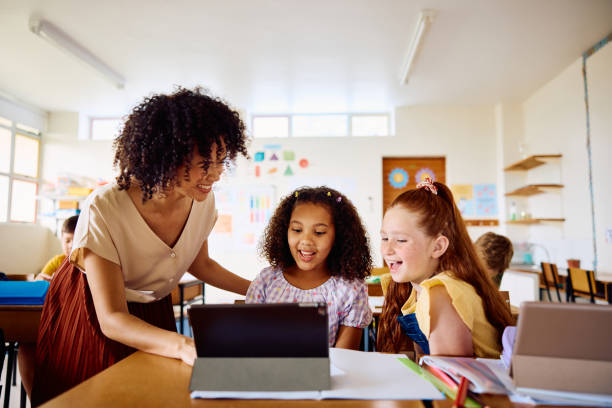 School teacher smiling to schoolgirls sitting at desk using digital tablet, explaining and laughing