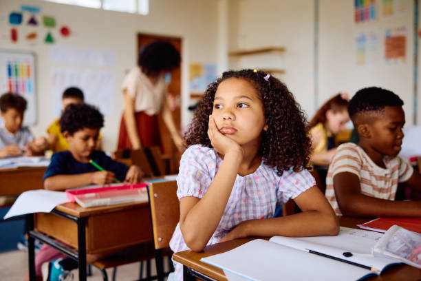 istock Elementary schoolgirl daydreaming at desk in class, looking sideways 2203067404