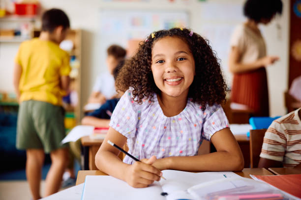 istock Elementary schoolgirl sitting at desk, smiling at camera in class 2203067413