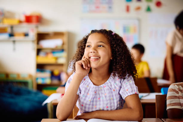 istock Elementary schoolgirl sitting at desk, looking away smiling in class 2203067464