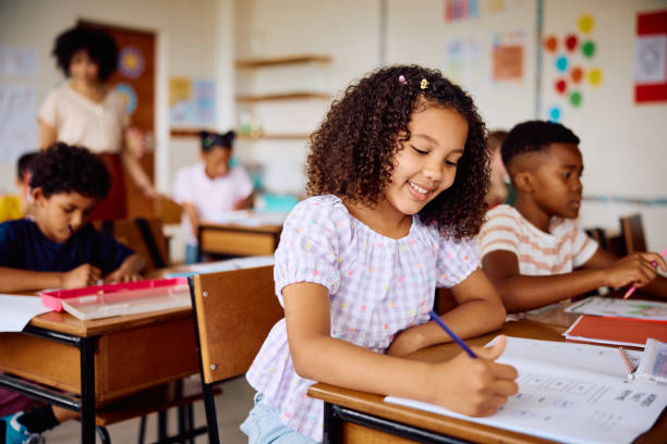 istock Elementary schoolgirl sitting at desk, smiling and writing in class 2203067480