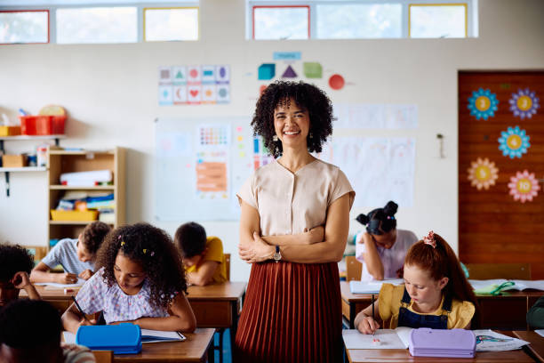istock Elementary school teacher smiling at camera with arms crossed in class 2203067562