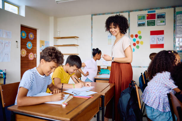 istock Elementary school teacher smiling at happy schoolboys writing at desk in classroom 2203067571
