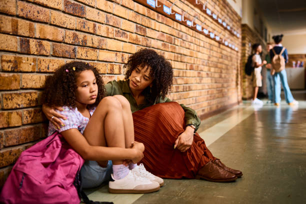 Elementary school teacher consoling schoolgirl leaning on wall looking sad in school corridor stock photo