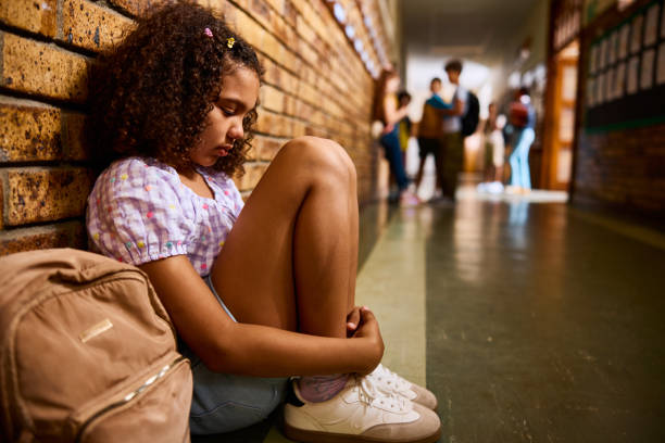 istock Schoolgirl sitting on floor, looking down sadly in school corridor, side view 2203067762