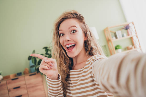 istock Young woman with blond hair smiling and pointing in striped pullover indoors 2203068005