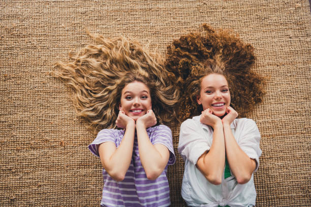 Two young women joyfully smiling while lying in a cozy interior. Embracing friendship and relaxation, they depict happiness and charming companionship in a delightful living room setting.