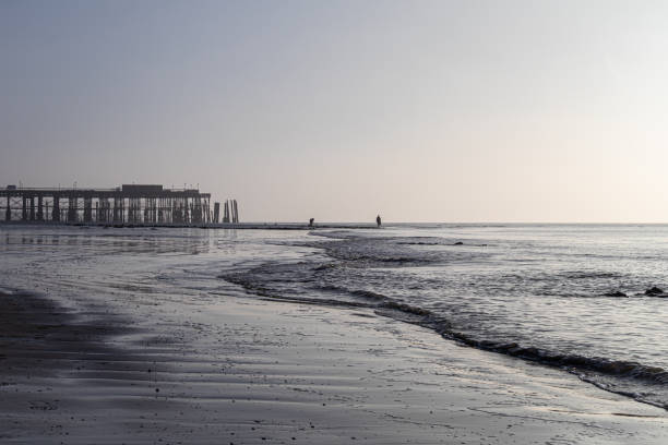 Hastings Pier reflected in the wet sand at low tide.  Two silhouetted people are in the scene, gentle waves roll across the sand