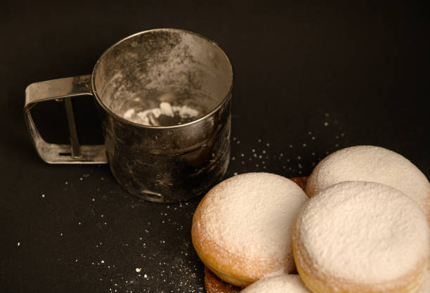 Golden-brown doughnuts dusted with powdered sugar beside a metal sifter on a dark background. Perfect for bakery branding, menu designs, or food-related content.