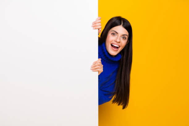 istock Young cheerful woman peeking from behind white panel against bright yellow backdrop wearing a blue sweater 2203078065