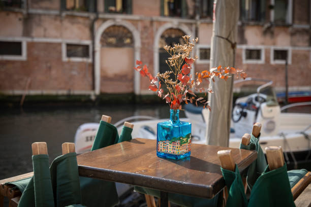 A blue vase made of glass filled with some twigs on a little table of a street restaurant at the edge of a small canal somewhere in Venice, Italy.\nCanon EOS R5 II, 1/1600, f/2,8 , 35 mm.