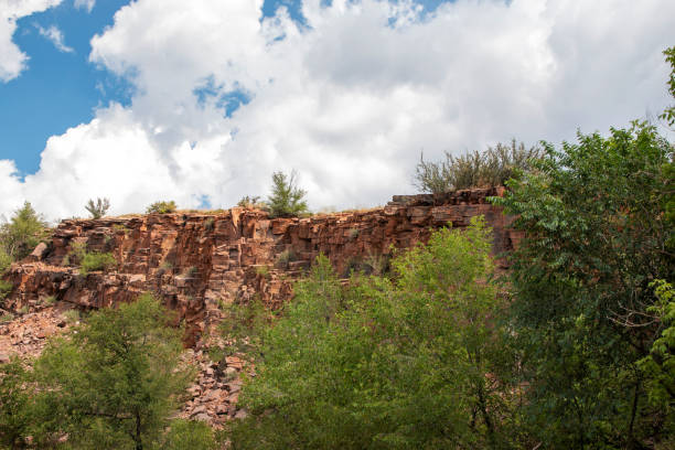 The rocky cliff rises majestically against a bright blue sky dotted with white clouds, surrounded by green trees.