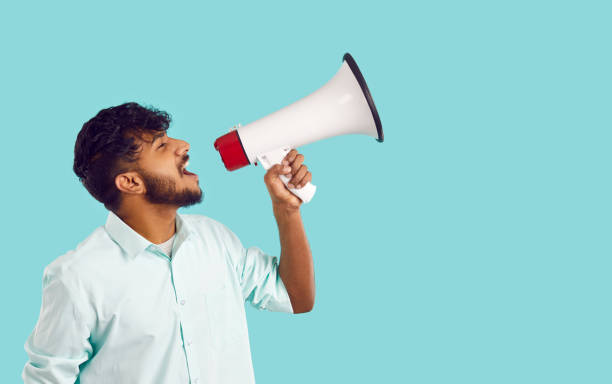 istock Young ethnic man with megaphone making loud marketing advertisement on light blue background. 2203092876