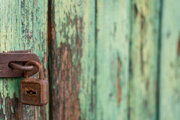 A heavily rusted padlock is attached to a rusty metal hasp on a weathered wooden surface. The wood features peeling green paint, revealing bare patches beneath, adding a rustic, aged look.
